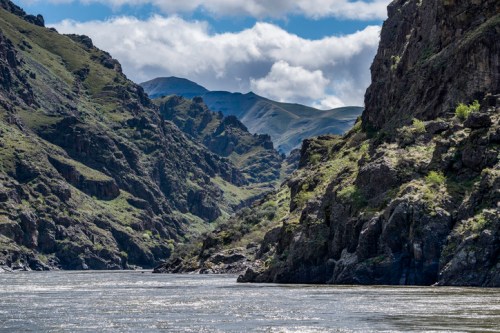 A serene river flows between steep, rocky cliffs under a partly cloudy sky, surrounded by lush green hills.