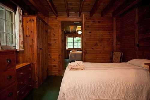 Cozy wooden cabin interior featuring a bed, dresser, and a doorway leading to another room. Natural light from windows.