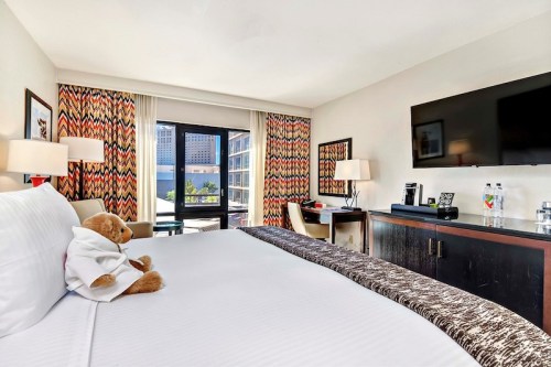 A cozy hotel room featuring a bed, a teddy bear in a robe, a TV, and colorful curtains with a view outside.