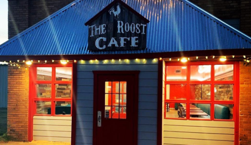 Exterior of The Roost Cafe featuring a red door, large windows, and a metal roof, illuminated by string lights.