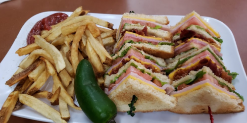 A plate of stacked sandwiches with ham, cheese, and lettuce, served with fries and a jalapeño pepper on the side.