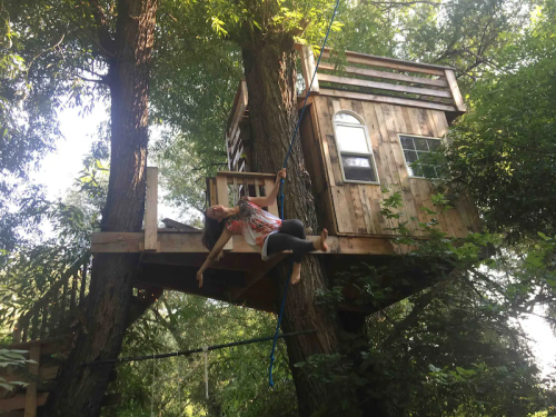 A person swings from a rope near a wooden treehouse nestled in lush green trees.