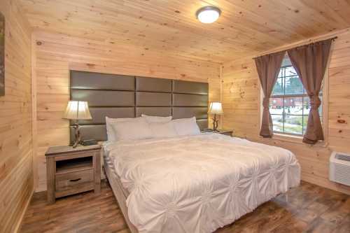 Cozy bedroom with a large bed, wooden walls, two lamps, and a window with curtains.