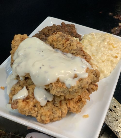 A plate of fried chicken topped with creamy gravy, served with sides of beans and macaroni salad.