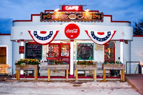 A charming café with patriotic decorations, featuring a Coca-Cola sign and flower boxes, set against a twilight sky.