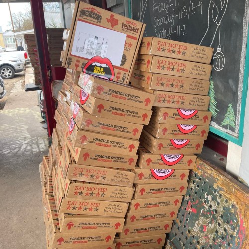 A stack of pizza boxes labeled "Eat Mo' Pie" outside a shop, with a chalkboard and delivery schedule in the background.
