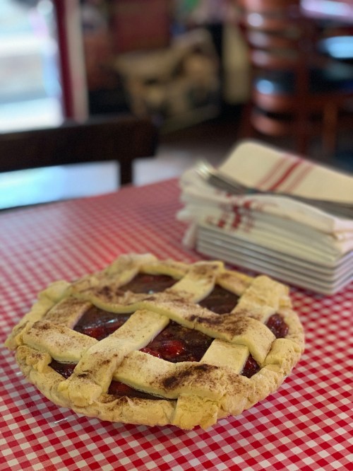 A freshly baked pie with a lattice crust sits on a red and white checkered tablecloth, next to stacked plates and napkins.