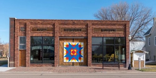 A brick building with large windows and a colorful quilt pattern mural on the door, set against a clear blue sky.