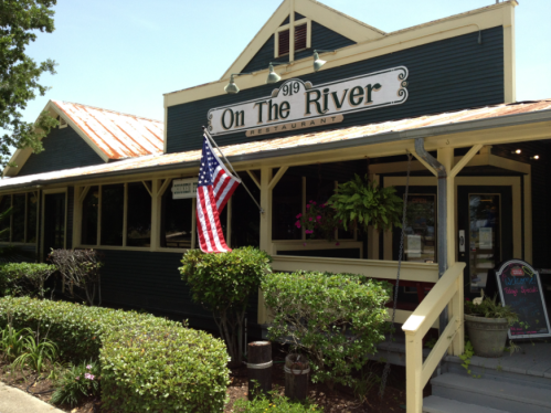 Exterior of "On The River" restaurant featuring a porch, American flag, and lush greenery.