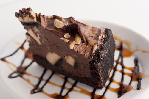 A slice of chocolate cheesecake with a cookie crust, topped with chocolate and nuts, drizzled with caramel sauce.