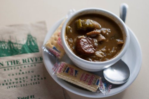 A bowl of gumbo with sausage and rice, served with crackers and a spoon, on a plate next to a restaurant napkin.