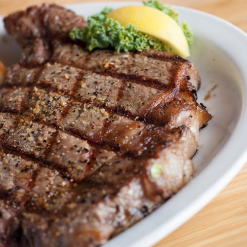 A perfectly grilled steak with a seared crust, garnished with a lemon wedge and fresh greens on a white plate.