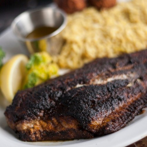 A close-up of a grilled fish fillet on a plate, served with rice, lemon, and a small cup of sauce.