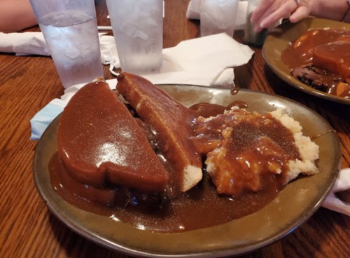 A plate of food featuring sliced meat with gravy, mashed potatoes, and bread, served with iced water in the background.