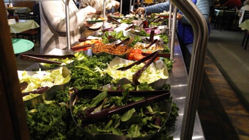 A vibrant salad bar with various greens, toppings, and utensils for serving, set in a dining area.