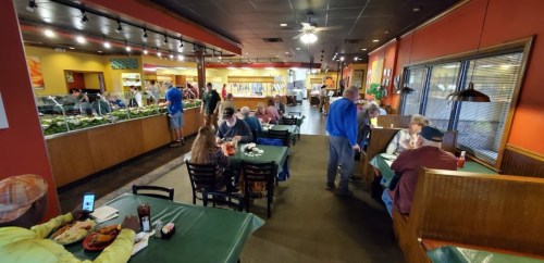 A busy restaurant interior with diners at tables and a buffet area in the background. Brightly colored walls and decor.