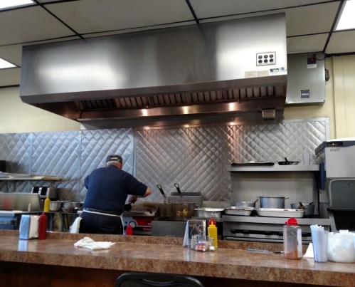A chef prepares food in a commercial kitchen, with stainless steel appliances and condiments on the counter.