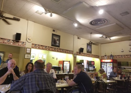 A casual diner scene with several people seated at tables, chatting, and a counter with drinks in the background.