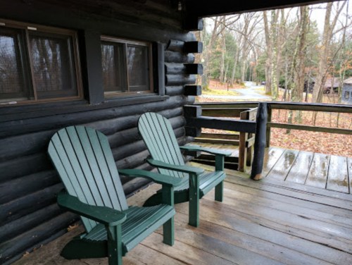Two green Adirondack chairs on a wooden porch, surrounded by trees and a winding path in the background.
