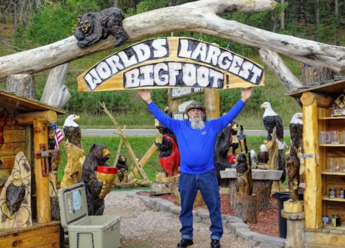 A man in a blue shirt stands under a sign reading "World's Largest Bigfoot," surrounded by wooden sculptures and nature.