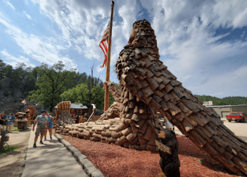 A large wooden sculpture of an eagle with people nearby, set against a cloudy sky and American flag.