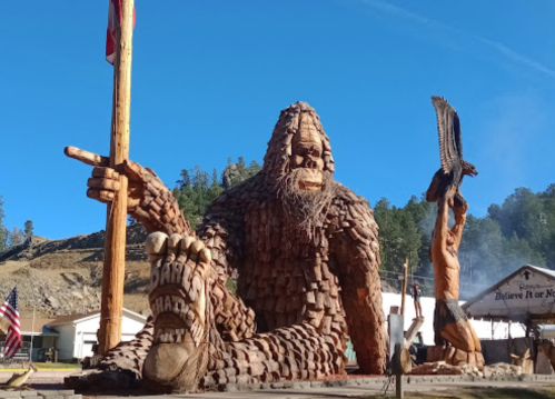 A large wooden sculpture of Bigfoot sits with a flag, alongside a smaller figure holding an eagle, set against a blue sky.