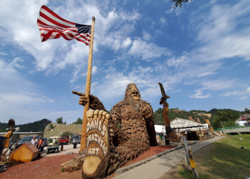 A large wooden Bigfoot statue holding an American flag, surrounded by trees and vehicles under a blue sky.