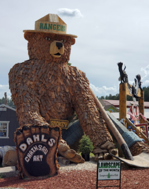 A large wooden bear sculpture wearing a ranger hat, with a sign reading "Dahl's Chainsaw Art" nearby.