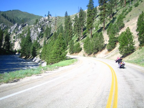 A motorcyclist rides along a winding road beside a river, surrounded by lush green trees and mountains.