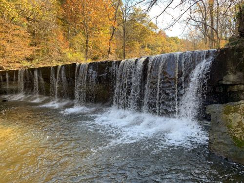 A serene waterfall cascades over a rocky ledge, surrounded by autumn foliage and a calm river.