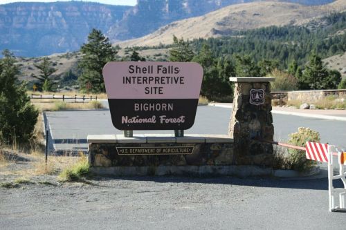 Sign for Shell Falls Interpretive Site in Bighorn National Forest, with mountains and trees in the background.