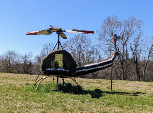 A whimsical helicopter sculpture made of hay and metal, set in a grassy field with trees in the background.