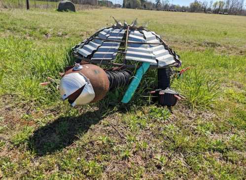 A whimsical turtle sculpture made from metal and found objects, resting in a grassy field under a clear blue sky.