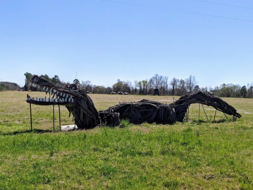 A large, artistic sculpture of a dragon made from branches, lying in a grassy field under a clear blue sky.