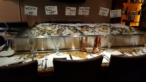 A seafood bar displaying oysters on ice with labels for different varieties and a set dining table nearby.