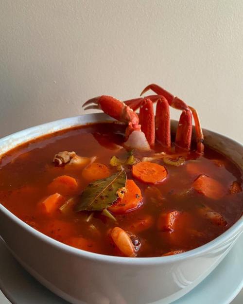 A bowl of rich, red soup with crab claws, carrots, and herbs, set against a plain background.