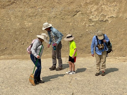 A group of four people, including two children, examining the ground in a dusty outdoor setting, wearing sun hats.
