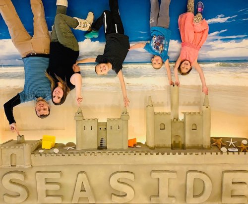 A family poses upside down above a sandcastle and the word "SEASIDE," with a beach backdrop.