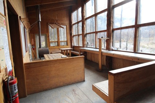 Interior of a rustic wooden building with large windows, featuring benches and a sink, surrounded by nature.