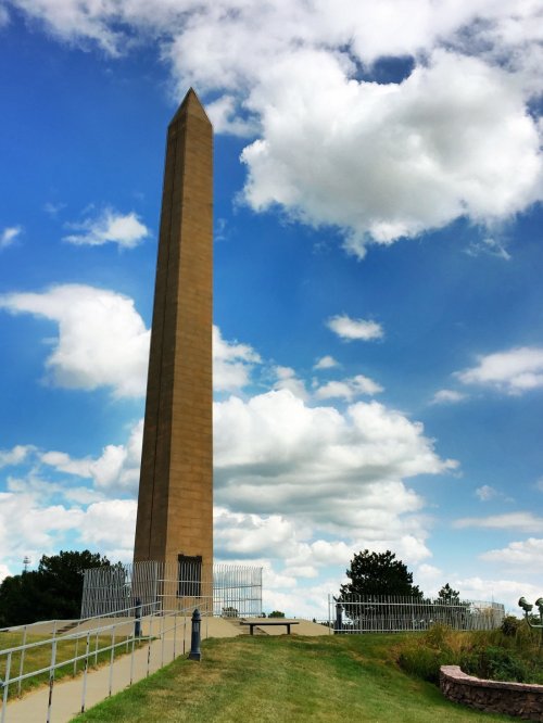 A tall obelisk stands against a blue sky with fluffy clouds, surrounded by green grass and trees.