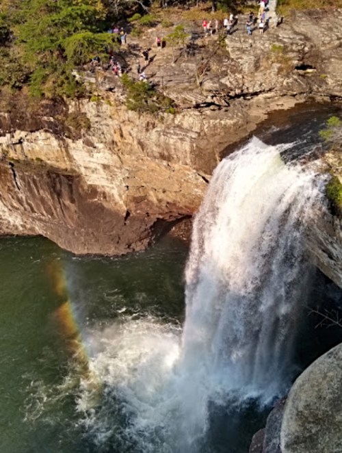 A waterfall cascades into a river, with a rainbow visible in the mist and people observing from the rocky ledge above.