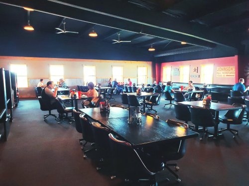A spacious restaurant interior with black tables and chairs, featuring diners and large windows letting in natural light.