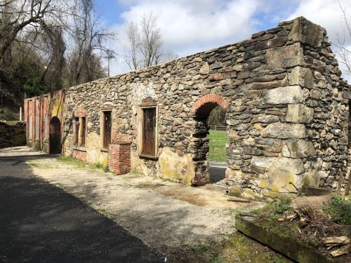 A weathered stone wall with arched openings, surrounded by trees and a gravel path under a partly cloudy sky.