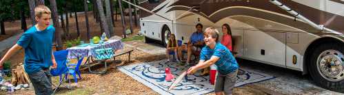 A family enjoys outdoor fun near an RV, with two boys playing a game while adults relax nearby.