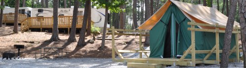 A green tent on a wooden platform in a forested campsite, with a trailer and picnic area in the background.