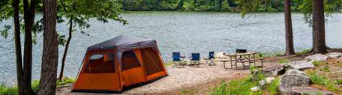 A bright orange tent by a calm lake, with camping chairs and a picnic table nearby, surrounded by trees.