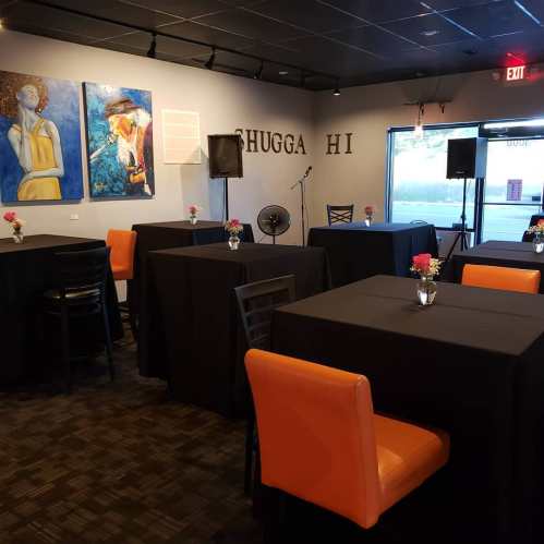 A cozy event space with black tables, orange chairs, and colorful artwork on the walls. Flowers on tables add a touch of decor.