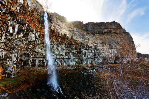 A stunning waterfall cascades down a rocky cliff, surrounded by autumn foliage and a clear blue sky.