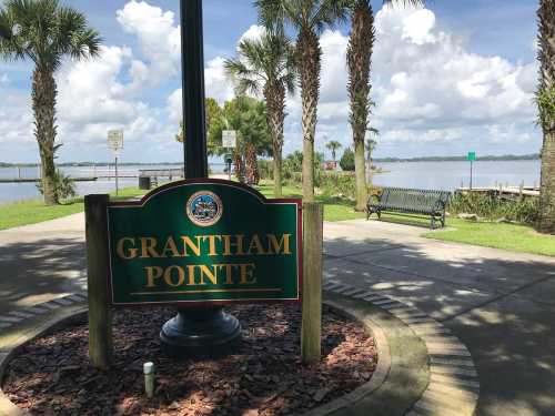 Sign for Grantham Pointe near a waterfront, surrounded by palm trees and benches under a partly cloudy sky.