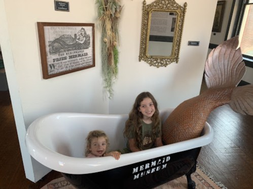 Two children sit in a vintage bathtub with a mermaid tail, surrounded by museum decor in a bright room.
